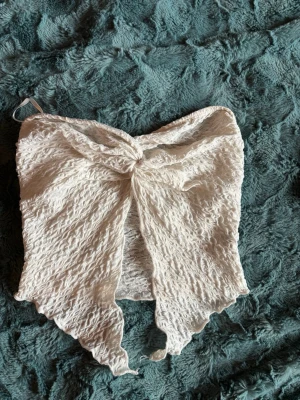 Vit bandeau tubtopp med knyt - Trendig vit bandeau tubtopp med knyt framtill och vågig kant. Toppen är gjord i ett strukturerat, lätt skrynkligt tyg som ger en cool look. Perfekt för att skapa en somrig och avslappnad stil.