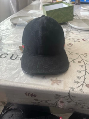 Svart keps från Gucci med GG-mönster - Snygg svart keps från Gucci med det klassiska GG-monogrammönstret över hela kepsen. Kepsen har en böjd skärm och justerbart band bak i läder. På sidan finns Guccis ikoniska gröna och röda rand. Perfekt accessoar för en trendig look.