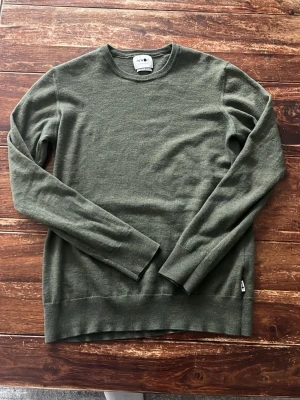 Olivgrön stickad tröja från NN07 - Snygg olivgrön stickad tröja från NN07 i mjukt italienskt garn. Storlek har varierats från L till Passform XS efter krympning men är fortfarande i jätte bra skick. Tröjan har rund halsringning, långa ärmar och en klassisk, stilren design som passar perfekt till jeans eller chinos. 