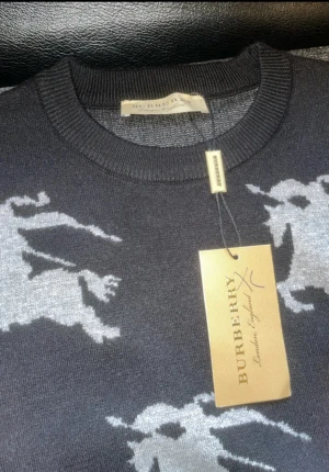 Helt ny burberry tröja - Snygg mörkgrå tröja från Burberry med ikoniskt ridande riddare-mönster i ljusgrått. Rund halsringning och ribbade muddar. Perfekt för dig som vill ha en stilren men ändå unik look. Materialet känns mjukt och bekvämt.