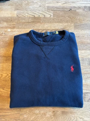 Mörkblå sweatshirt från Polo Ralph Lauren - Mörkblå sweatshirt från Polo Ralph Lauren med klassisk rund halsringning och röd broderad logga på bröstet. Tröjan är i mjukt material och har en avslappnad passform, perfekt för en stilren och bekväm look.