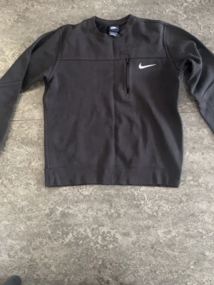 Svart sweatshirt från Nike - Svart sweatshirt från Nike med rund halsringning och en diskret bröstficka med dragkedja. Tröjan har lång ärm och klassisk Nike-logga i vitt på bröstet. Perfekt för en sportig och avslappnad stil.