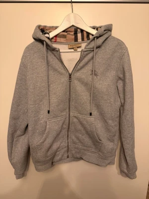 Grå Burberry kofta - Säljer en grå hoodie från Burberry med klassiskt rutigt foder i huvan och dragkedja framtill. Hoodien har två stora fickor, snören i huvan och broderad logga på bröstet. Materialet är mjukt och bekvämt, perfekt för chill dagar.