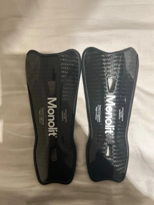 Svarta benskydd från Monolit - Säljer ett par svarta benskydd från Monolit med vit logga. De har en lätt böjd form och två hål för ventilation. Ytan har ett mönster som liknar kolfiber och skydden är tillverkade i ett hårt syntetmaterial. Perfekta för fotboll eller andra sporter.