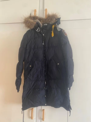 Mörkblå dunjacka från Parajumpers - Snygg mörkblå dunjacka från Parajumpers med längre modell, huva med äkta päls och gul detalj vid halsen. Jackan har två stora fickor framtill och patch på axeln. Perfekt för kalla vinterdagar.