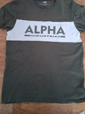 Grön Alpha Industries t-shirt - Säljer en grön t-shirt från Alpha Industries med vit panel och logotyp på bröstet. T-shirten har rund hals och korta ärmar, perfekt för dig som gillar streetwear och vill ha en enkel men snygg look.