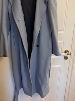 Mango lång kappa - Snygg ljusblå lång rock med bälte i midjan och klassisk krage. Rocken har en enkel knäppning framtill och är tillverkad i ett mjukt tyg som ger en stilren look. Perfekt för att höja din outfit under kalla dagar.