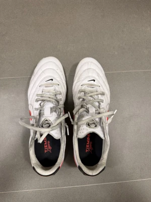 Nike Tiempo Ligera FG storlek 43 - Säljer ett par vita Nike Tiempo Legend fotbollsskor med svart och röd swoosh på sidan. Skorna har klassisk snörning, låg profil och dobbar för spel på gräs. Ovandelen är i mjukt läder för bra bollkontroll och komfort. Perfekta för dig som vill ha stilrena och funktionella skor.