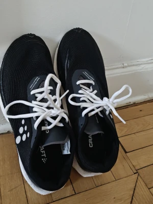 Svarta Craft sneakers med vita detaljer 43,5 - Snygga svarta sneakers från Craft med vita snören och vit under sula samt svarta inre sulor. Skorna har en sportig design med diskreta vita prickar på sidan och är tillverkade i ett lätt och ventilerande material. Perfekta för dig som gillar en stilren och modern look. Skorna är väl skötta, sparsamt använda och kommer från rökfritt och djurfritt hem. Skorna är 43,5 men rekommenderas bäst till 42,5 och 43 storlek havare. Förfrågor släng gärna ett mail.  