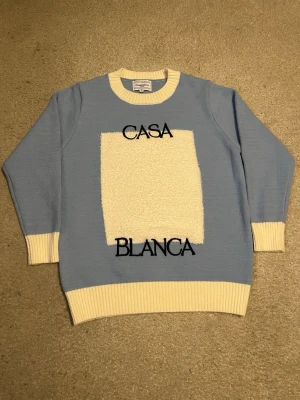 Blå stickad tröja från Casablanca - Ljusblå stickad tröja från Casablanca med kontrasterande vita muddar och krage. Framsidan har en vit, fluffig ruta och svart broderad text 'CASA BLANCA'. Perfekt för dig som gillar unik design och streetstyle.