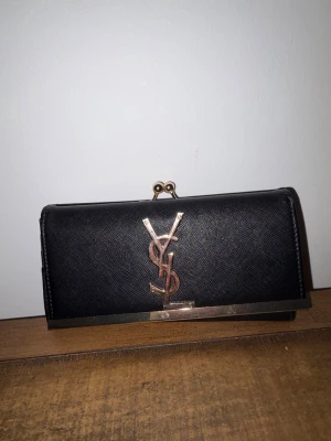 Svart YSL plånbok med gulddetaljer - Snygg svart plånbok från Turkiet med klassisk YSL-logga i guld framtill. Plånboken är tillverkad i strukturerat läder och har metallram med tryckknappsstängning. Insidan har flera fack för kort och sedlar. Perfekt accessoar för dig som gillar lyxiga detaljer.