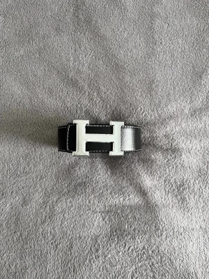 Herme’s bälte  - Snyggt svart skärp med ett stort, silverfärgat H-spänne framtill. Skärpet har vita sömmar längs kanterna och är gjort i slätt skinn. Perfekt accessoar för att lyfta vilken outfit som helst.