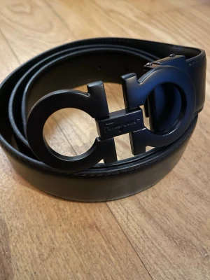 Svart skärp från Salvatore Ferragamo - Stilrent svart skärp från Salvatore Ferragamo med ikoniskt dubbel-Gancini spänne i matt svart finish.  har en klassisk, tidlös design som passar perfekt till jeans eller kostymbyxor.