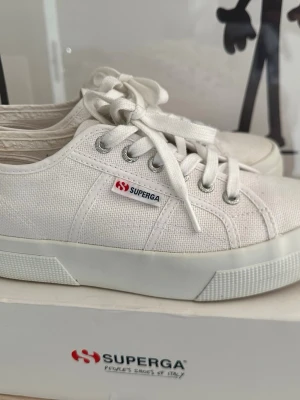 Vita Superga canvas sneakers - Klassiska vita sneakers från Superga i canvas med lågt skaft och vita snören. Skorna har en vit gummisula med lite platå och metallfärgade öljetter. Perfekta för en clean och stilren look. Strl 38. Använda 3ggr. Nypris 999:-
