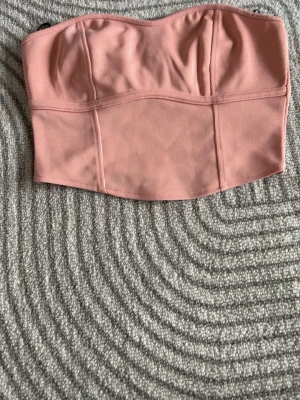Rosa bandeau tubtopp från H&M - Säljer en snygg rosa bandeau tubtopp från H&M. Toppen är axelbandslös och har en enkel, stilren design med markerade sömmar framtill. Perfekt att matcha med höga jeans eller kjol för en trendig look.