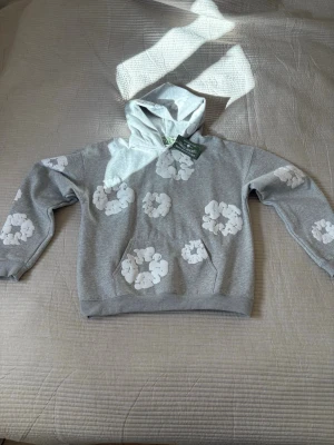 Grå hoodie från Denim Tears med blommor - Säljer en grå hoodie från Denim Tears med vita blommönster över hela tröjan. Hoodien har en klassisk huva och magficka framtill. Materialet är mjukt och bekvämt, perfekt för en avslappnad stil. Snygg detalj med märkets logga på etiketten.