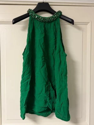 Grön ärmlös topp med pärldetaljer - Snygg grön ärmlös topp med hög hals och glittrande pärldetaljer runt kragen. Perfekt för dig som vill sticka ut med en färgstark och elegant look. Luftigt material och loose passform.
