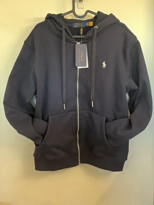 Polo Ralph Lauren hoodie - Snygg mörkblå hoodie från Polo Ralph Lauren med dragkedja, huva med dragsko och två stora fickor framtill. Klassisk logga broderad på bröstet. Tillverkad i mjukt bomullsmaterial som känns riktigt skönt mot huden. Perfekt för en avslappnad och stilren look.