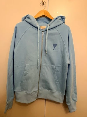 Ljusblå hoodie med dragkedja Ami - Ljusblå hoodie från Ami med dragkedja framtill, huva med dragsko och kängurufickor. Broderad Ami-logga med hjärta på bröstet. Mjuk bomullskänsla och avslappnad passform, perfekt för en chill stil.