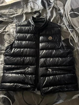 Moncler gui  - Snygg marinblå dunväst från Moncler med glansig finish och klassisk logga på bröstet. Västen har två dragkedjefickor framtill och stängs med dragkedja. Perfekt för lager-på-lager och håller dig varm med stil.