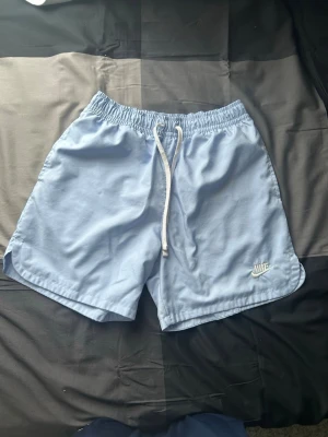 Ljusblå badbyxor från Nike - Snygga ljusblå badbyxor från Nike med vit dragsko i midjan och elastisk resår. Klassisk passform med meshfoder på insidan och diskret Nike-logga broderad på vänster ben. Perfekta för stranden eller poolen.