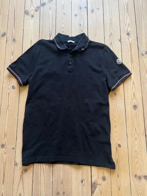 Svart Moncler pikétröja - Svart pikétröja från Moncler med klassisk krage, knappar framtill och logotyp på ärmen. Tröjan har ribbade muddar med tunna röda och vita ränder samt är tillverkad i bomull. Perfekt för en clean och stilren look. Storlek xxl men passar snarare l. Priset kan diskuteras och skriv gärna till mig om funderingar kring tröjan!
