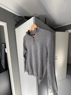 Grå halvzip stickad tröja - Snygg grå stickad tröja från Oscar Jacobson med halv dragkedja vid halsen. Tröjan har lång ärm och ribbade muddar vid ärmslut och nederkant. Perfekt för lager-på-lager och enkel att matcha med jeans eller chinos.