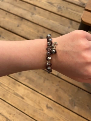 Lila pärlarmband med silverdetaljer - Säljer ett snyggt armband med lila och silverfärgade pärlor. Armbandet har en elastisk design och en liten silverfärgad berlock som detalj. Perfekt för att ge din outfit lite extra glans och stil.