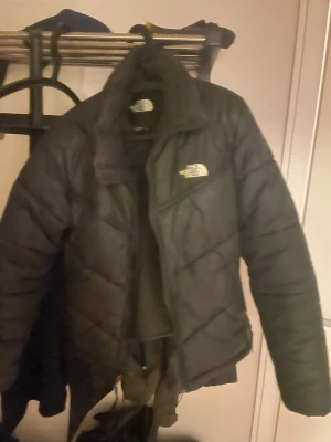 Svart pufferjacka från The North Face - Säljer en svart pufferjacka från The North Face med hög krage och dragkedja. Jackan har en klassisk quiltad design och logga på bröstet och ärmen. Perfekt för kalla vinterdagar och riktigt skön att bära. Materialet är slitstarkt och håller värmen bra.