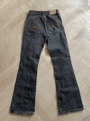 Svartgråa bootcut jeans från Nudie Jeans, Flare Glenn - Säljer ett par svarta bootcut jeans från Nudie Jeans med klassiska femficksdesign och orangea sömmar på bakfickorna. Jeansen har rå kant längst ner och är tillverkade i denim. Perfekt för dig som gillar en avslappnad och cool stil.