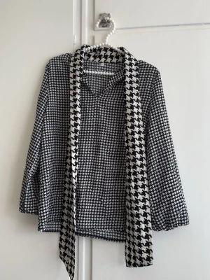 Svartvit blus med scarfdetalj - Snygg svartvit blus med klassiskt hundtandsmönster och matchande scarfdetalj i större mönster. Blusen har krage och långa ärmar med lätt puff vid ärmslut. Perfekt för dig som gillar retrovibbar och vill sticka ut lite extra.