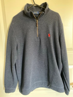 Mörkblå ziptröja från Polo Ralph Lauren - Mörkblå stickad ziptröja från Polo Ralph Lauren med klassisk röd broderad logga på bröstet. Tröjan har hög krage, dragkedja med läderdetalj och ribbade muddar. Perfekt för en stilren och avslappnad look.