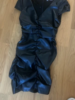 Svart omlottklänning från Zara - Snygg svart klänning från Zara i omlottmodell med kort ärm och coola rynkade detaljer längs hela framsidan. Klänningen har en glansig finish och är tillverkad i syntetmaterial. Perfekt för dig som vill sticka ut med en trendig look.