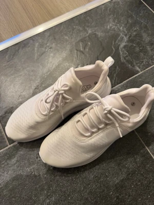 Helvita sneakers med sportig look - Snygga helvita sneakers med modern och sportig design. Skorna har en låg siluett, rund tå och ovandel i textil med diskreta mönsterdetaljer. Sulan är vit och ger bra grepp. Perfekta för dig som gillar en clean och stilren stil.