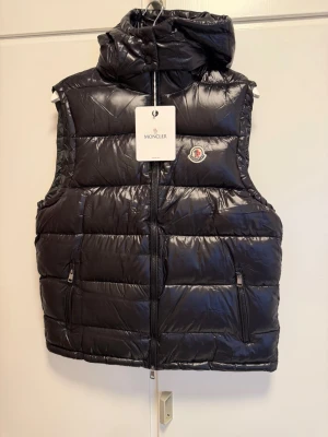 Svart dunväst - Snygg svart dunväst med glansig finish och ikonisk logga på bröstet. Västen har huva, två dragkedjeförsedda fickor och quiltad design. Perfekt för lager-på-lager och streetstyle. Materialet är dunfyllning och yttertyget är syntet. 