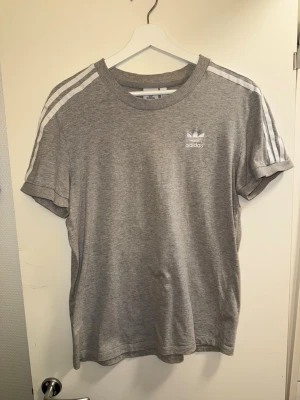 Grå Adidas t-shirt med vita ränder - Säljer en grå t-shirt från Adidas med klassiska vita ränder på axlarna och Adidas-logga på bröstet. T-shirten har rund hals och korta ärmar, perfekt för en avslappnad och sportig look. Materialet är mjukt och känns skönt mot huden.