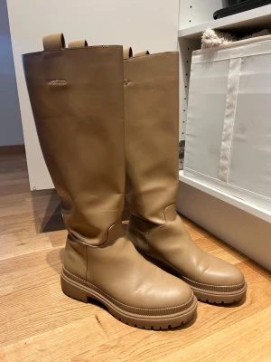 Beige höga stövlar i läder - Säljer ett par beige höga stövlar med chunky sula och dragflikar upptill. Stövlarna är tillverkade i slätt fejk läder och har en stilren, minimalistisk design utan synliga detaljer eller mönster. Perfekta för att lyfta din outfit med en modern touch.