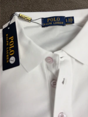 Vit pikéskjorta från Polo Ralph Lauren - Vit pikéskjorta från Polo Ralph Lauren med klassisk krage och knappar framtill. Skjortan har korta ärmar och den ikoniska logotypen broderad på bröstet. Perfekt för dig som gillar stilrena och tidlösa plagg.