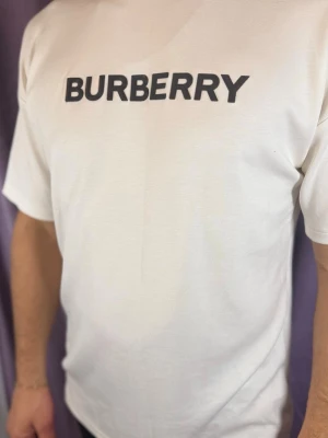 Vit Burberry t-shirt med logotyp - Säljer en vit t-shirt från Burberry med klassisk svart logotyp på bröstet. T-shirten har en rak passform och korta ärmar, perfekt för dig som gillar stilrena plagg med exklusiv känsla. Materialet är mjuk bomull som känns skönt mot huden.