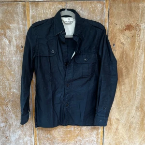Svart overshirt från Beauty & Youth - Stilren svart overshirt från Beauty & Youth United Arrows. Skjortan har klassisk krage, två bröstfickor med knappar och långa ärmar. Perfekt att bära öppen eller stängd för en avslappnad look. Gjord i bomull för skön känsla.