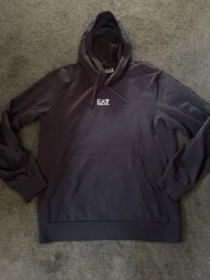 Svart hoodie från Emporio Armani EA7 - Snygg svart hoodie från Emporio Armani EA7 med vit logga på bröstet. Klassisk modell med huva och dragsnören samt stor magficka framtill. Perfekt för en avslappnad och trendig streetwear-look.