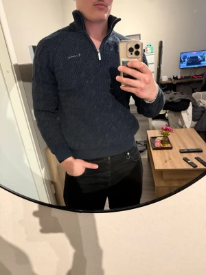 Mörkblå stickad tröja med dragkedja - Mörkblå stickad tröja från Lindbergh med halv dragkedja och hög krage. Tröjan har ett snyggt strukturmönster och broderad logga på bröstet. Perfekt för lager-på-lager och passar till både jeans och chinos.