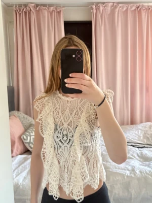 Vit spetsblus med volangdetaljer - Supersöt vit blus i spets med volangdetaljer och hög hals. Ärmlös modell med vackert mönstrad spets och scallop-kant nertill. Perfekt för dig som vill ha en romantisk och trendig topp till sommaren.