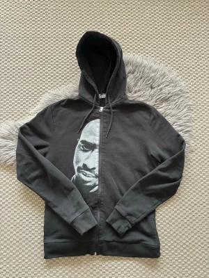 Svart hoodie med tryck av Tupac Shakur - Svart hoodie med dragkedja och huva, prydd med ett stort tryck av ett 2Pack på framsidan. Hoodien har långa ärmar, ribbade muddar och dragsko i huvan. Perfekt för en avslappnad streetwear-look.