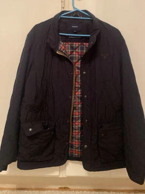 Svart quiltad jacka från Gant - Snygg svart quiltad jacka från Gant med rutigt innerfoder i rött, vitt och blått. Jackan har ståkrage, dragkedja och knappar framtill samt två stora fickor med lock. Perfekt för kyliga dagar och ger en klassisk look.