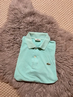 Mintgrön pikétröja från Lacoste - Snygg mintgrön pikétröja från Lacoste med klassisk krage och två knappar framtill. Tröjan har korta ärmar och den ikoniska krokodilloggan broderad på bröstet. Perfekt för en avslappnad och stilren look.