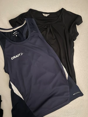 Träningstopp från Craft & svart topp - Två toppar: en marinblå sportig linnetopp från Craft med vita detaljer, perfekt för träning, och en enkel svart kortärmad topp med rund halsringning. Båda är i mjukt, syntetiskt material som andas. Marinblå tröja är i storlek S men är mycket mindre i verkligheten 