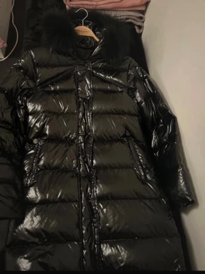 Glansig svart dunjacka med pälskrage, från Garoff - Snygg svart lång dunjacka med glansig finish och stor fluffig pälskrage på huvan. Jackan har dragkedja och knappar framtill samt två fickor. Perfekt för kalla vinterdagar och riktigt varm.