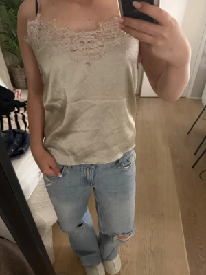 Champagnefärgad linnetopp Twist & Tango - Champagnefärgad linnetopp från Twist & Tango med tunna axelband och vacker spetsdetalj vid urringningen. Sidenliknande material som ger en lyxig känsla. Perfekt att styla med jeans eller kjol för en trendig look.