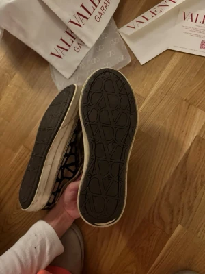 Valentino Garavani sneakers - Snygga svarta sneakers från Valentino Garavani med vit sula och snörning. Skorna har ett grafiskt mönster med Valentino-loggor över hela ovandelen och klassisk rund tå. Skorna är i riktigt bra skick och allt OG ingår, tveka inte på att höra av er vid funderingar 